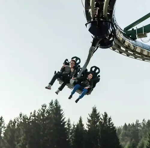 Zwei Personen schweben mit dem Wie-Flyer der Wiegand Erlebnisberge hoch über den Baumwipfeln. Der Betreiber von Sommerrodelbahnen bietet mit dieser Attraktion ein einzigartiges Flugerlebnis in luftiger Höhe