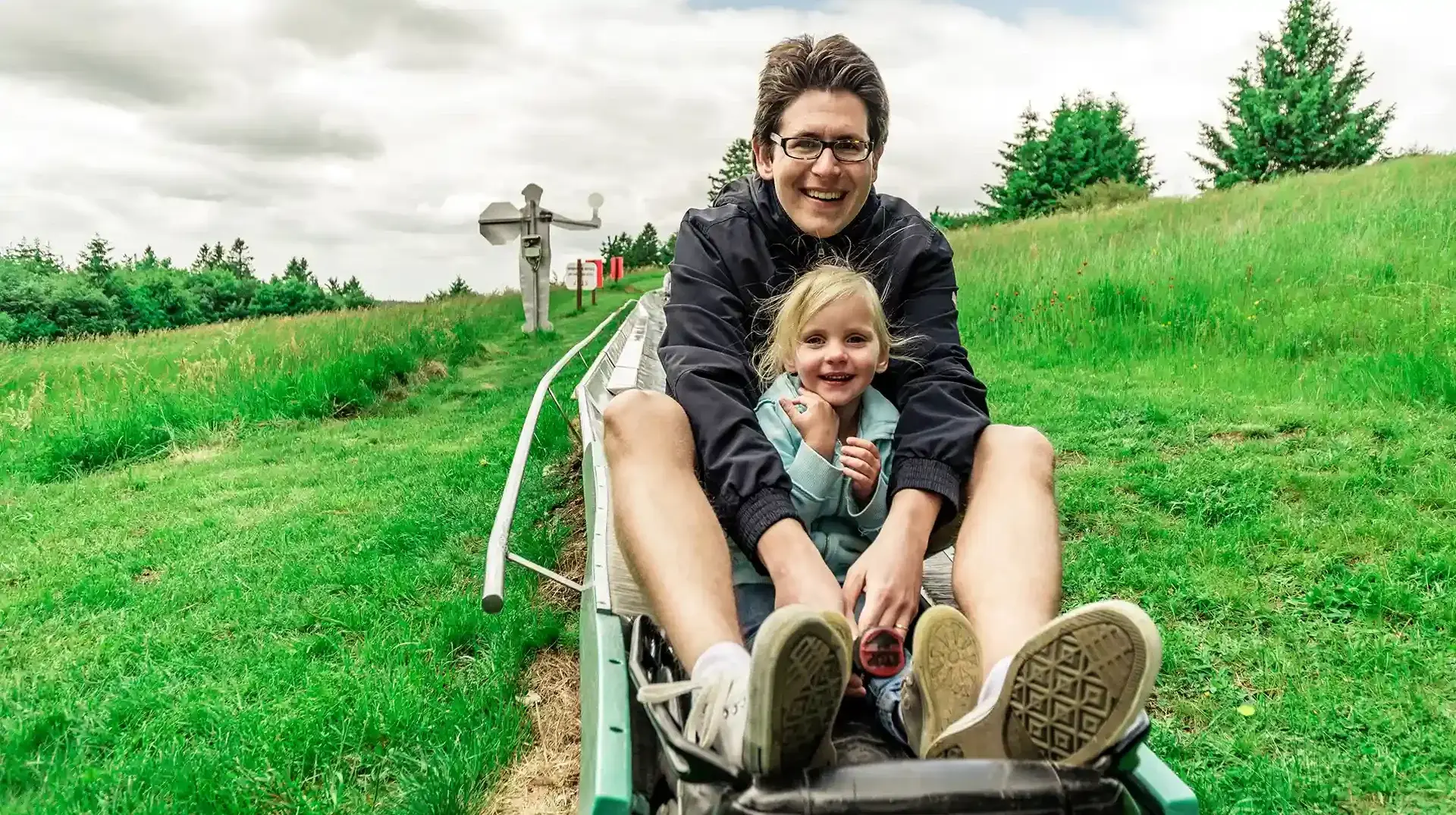 Eine frau und ihre Tochter fahren zusammen mit der Sommerrodelbahn. Die Mutter hält ihre Tochter fest und hat den Bremshebel in der Hand. Im Hintergrund sieht man Wiesen sowie Bäume. 