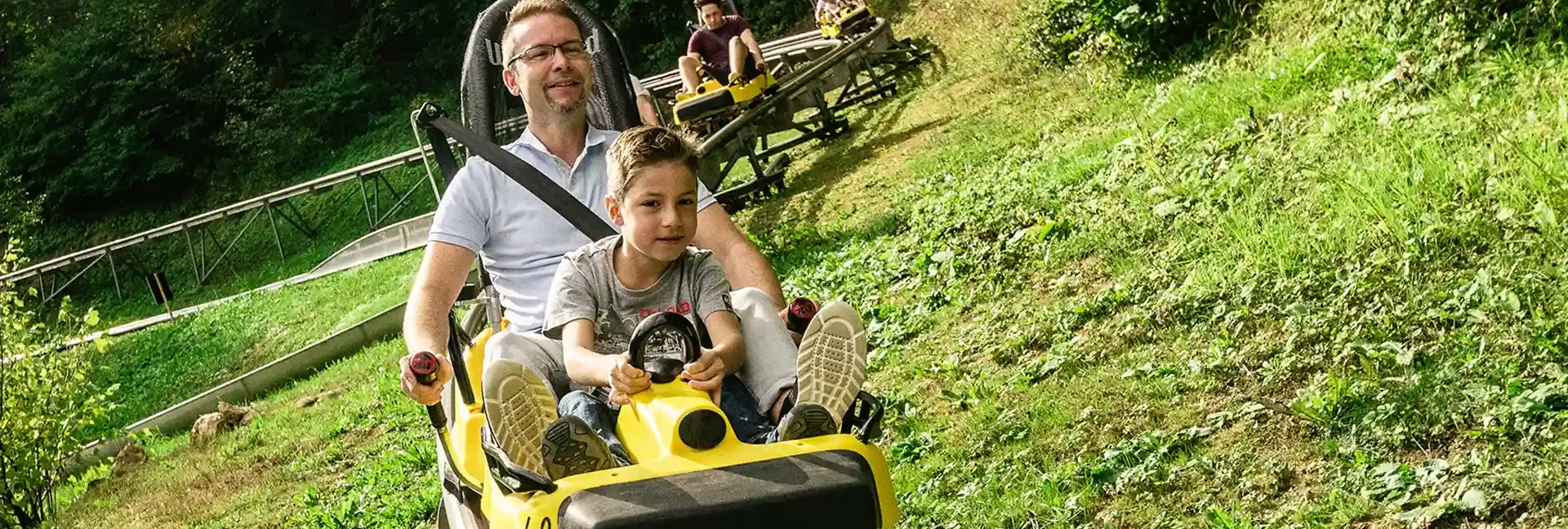 Ein Vater und sein Sohn fahren mit der Sommerrodelbahn. Beide sind am lachen. Im Hintergrund sieht man noch andere Fahrer auf der Sommerrodelbahn sowie Wiesen und Bäume.