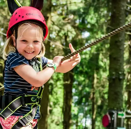 Ein lachendes Kind mit Klettergurt und Helm erklimmt den Kletterwald der Wiegand Erlebnisberge. Der Betreiber von Sommerrodelbahnen legt großen Wert auf Sicherheit und Abenteuer für die ganze Familie