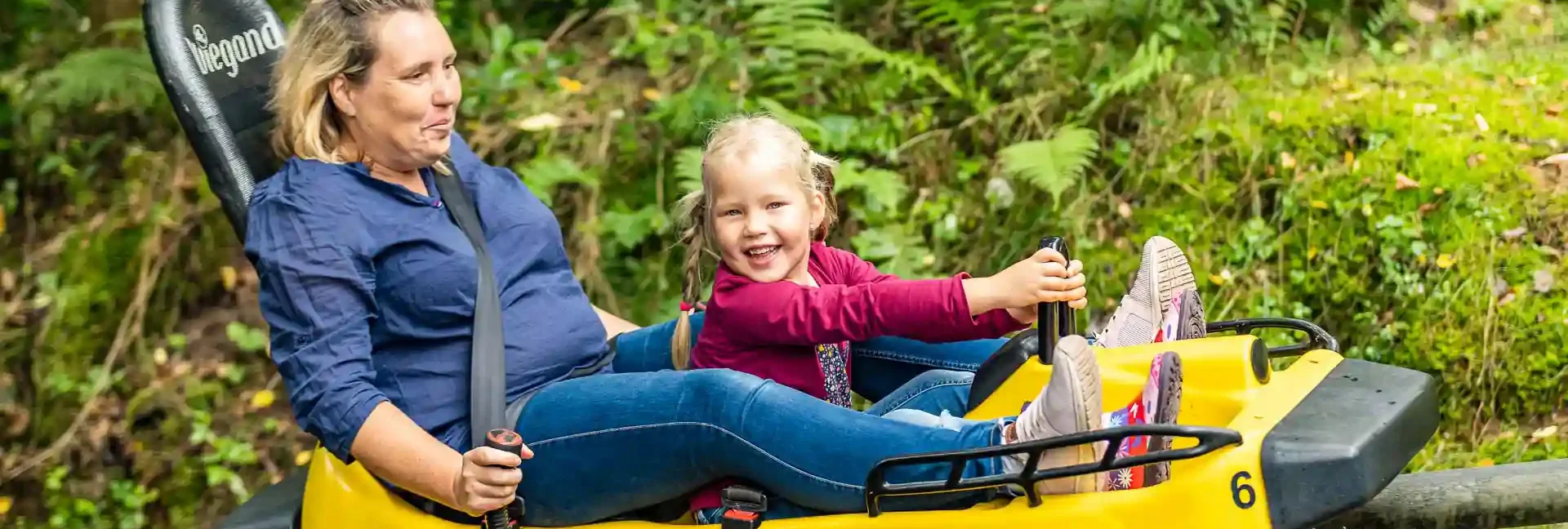 Eine Mutter und ihre Tochter fahren zusammen mit der Sommerrodelbahn. Die Tochter hält sich fest und die Mutter hat die Bremshebel in der Hand. Die Tochter ist am lachen und die Mutter schaut auch sehr engagiert. Im Hintergrund sind Bäume und Wiesen zu sehen.