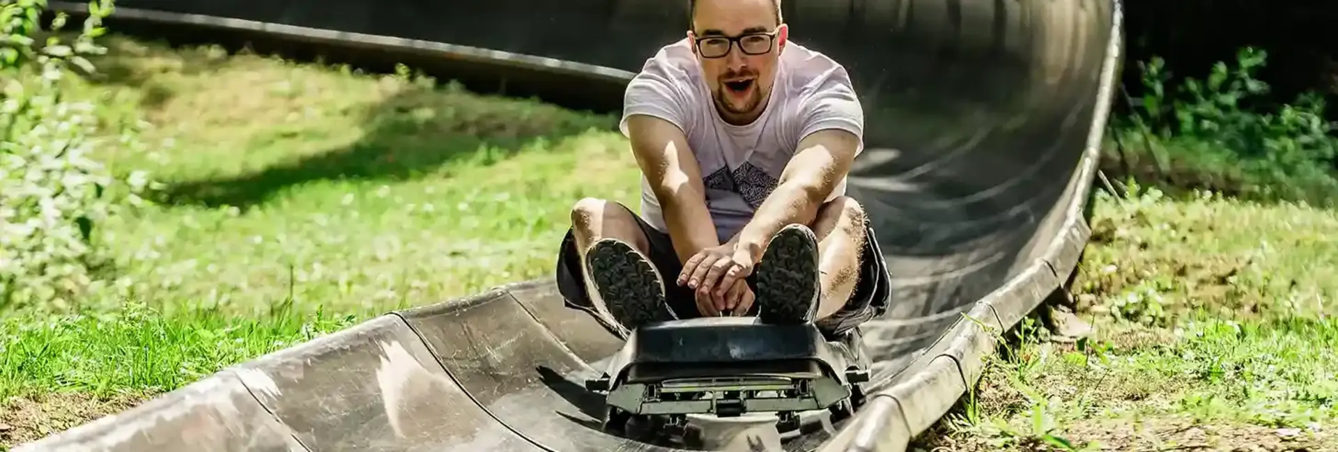 Ein Mann fährt im Sommer auf einer Sommerrodelbahn. Er fährt inmitten der Natur und hat sichtlich Spaß. Er hält sich am Schlitten fest.