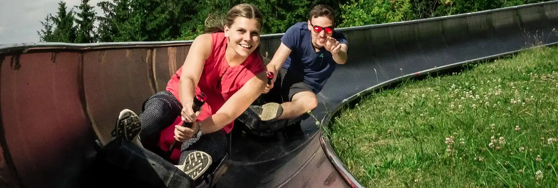 Eine Frau fährt mit der Sommerrodelbahn. Hinter ihr fährt ihr Freund auf einem einzelnen Schlitten. Der Mann zeigt in die Kamera und beide sind am lachen. Im Hintergrund der Bahn sieht man Bäume und Wiese.