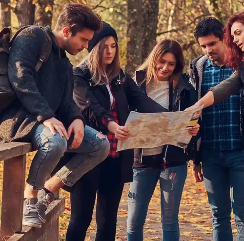 Eine Gruppe junger Erwachsener ist auf einer Brücke. Die hälfte von ihnen sitzt auf dem Geländer der Brücke, die anderen stehen. Sie schauen sich zusammen eine Karte an. Die Brücke liegt innerhalb eines Waldstückes.