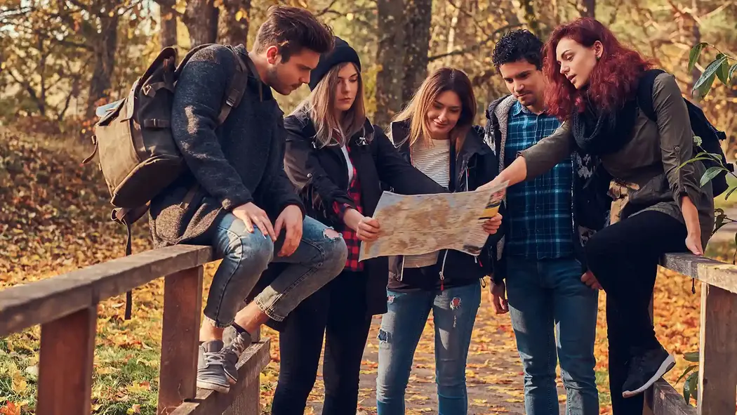 Eine Gruppe junger Erwachsener ist auf einer Brücke. Die hälfte von ihnen sitzt auf dem Geländer der Brücke, die anderen stehen. Sie schauen sich zusammen eine Karte an. Die Brücke liegt innerhalb eines Waldstückes.