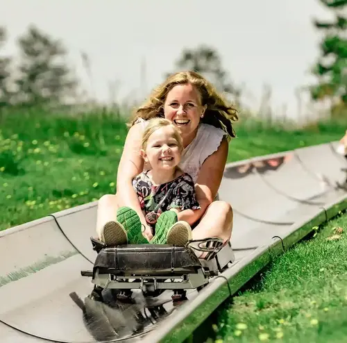 Eine Mutter und ihr Kind fahren lachend die Sommerrodelbahn der Wiegand Erlebnisberge hinunter. Der Betreiber von Sommerrodelbahnen sorgt für actionreichen Freizeitspaß in grüner Umgebung