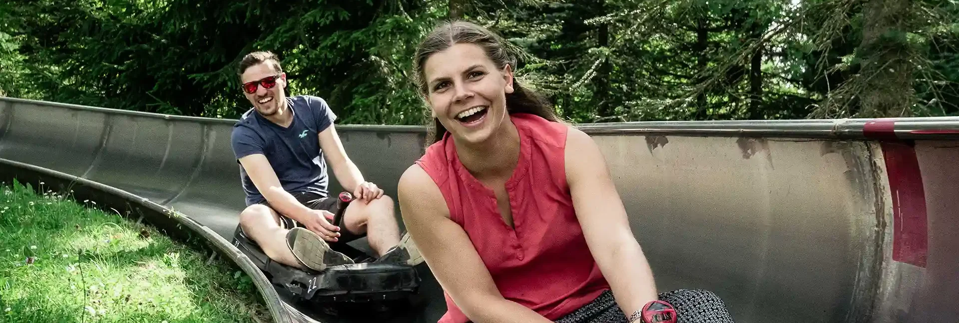 Zwei junge Erwachsene fahren lachend mit der Sommerrodelbahn. Im Hintergrund sieht man Bäume und neben der Bahn fängt direkt die Wiese an.