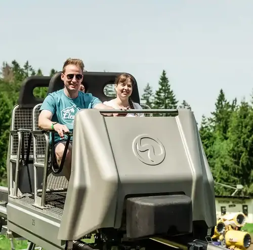 Ein Paar fährt mit dem Wie-Li der Wiegand Erlebnisberge, dem Betreiber von Sommerrodelbahnen, über eine Schienenstrecke durch die grüne Landschaft