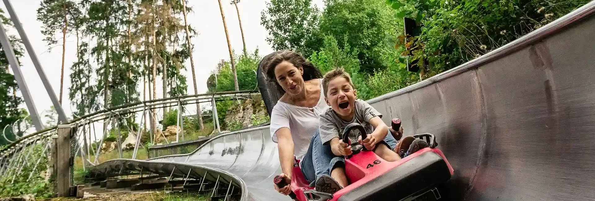 Eine Frau mit ihrem Sohn fahren zusammen mit der Sommerrodelbahn durch die grüne Natur. Der Junge hält sich fest und die Frau hat den Bremshebel in der Hand um die Geschwindigkeit zu steuern. Beide haben großen Spaß. Entdecken sie spannende Attraktionen an den Wiegand Erlebnisbergen.