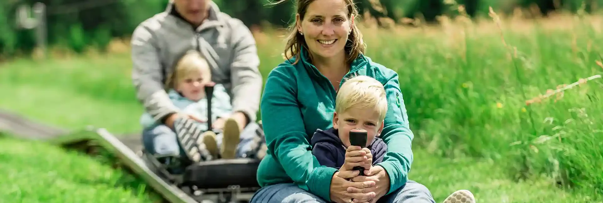 Eine Mutter und Ihr Sohn fahren mit der Sommerrodelbahn. Ihr Mann und die Tochter fahren in einem eigenen Schlitten dahinter. Sie fahren durch die grüne Natur.