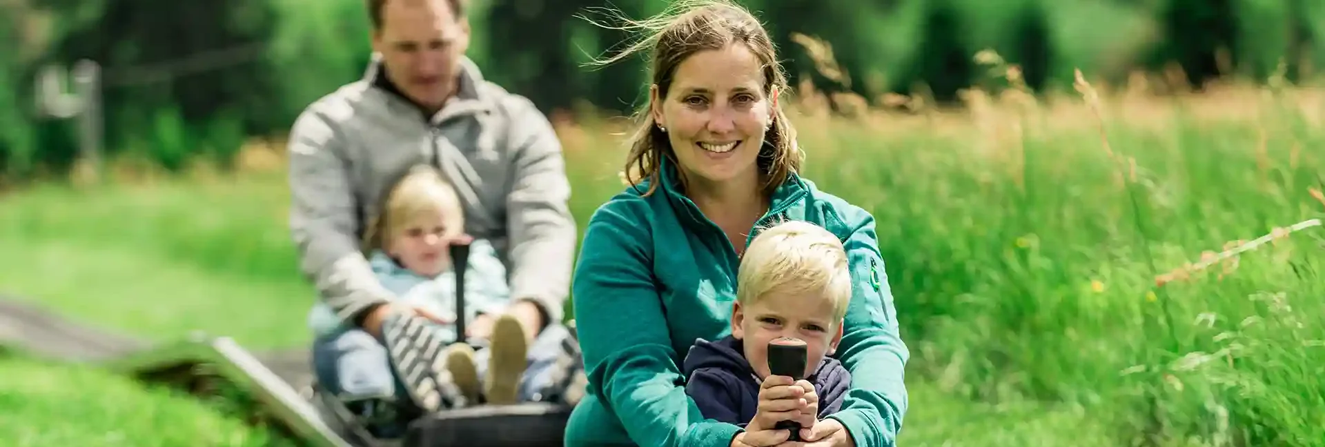 Eine Mutter und Ihr Sohn fahren mit der Sommerrodelbahn. Ihr Mann und die Tochter fahren in einem eigenen Schlitten dahinter. Sie fahren durch die grüne Natur.