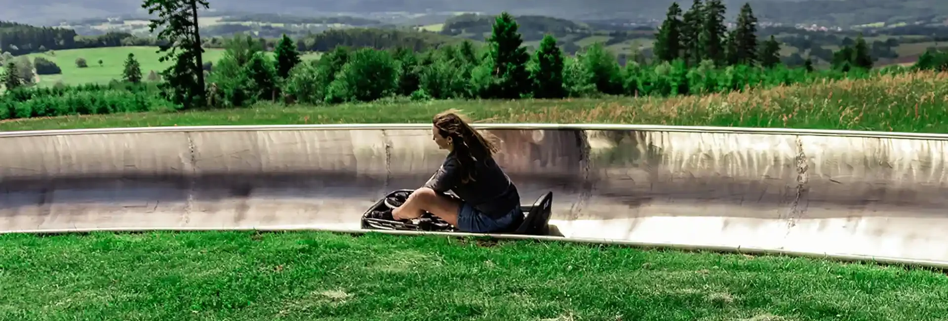Man sieht eine junge Frau auf der Sommerrodelbahn. Sie fährt durch eine Kurve und im Hintergrund sieht man die Natur, Bäume und Wiesen.